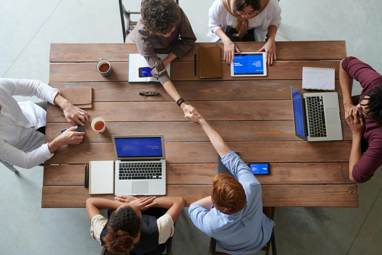 Personen sitzen an einem Arbeitstisch mit Laptops und führen eine Besprechung. Zwei Personen geben sich die Hand. 