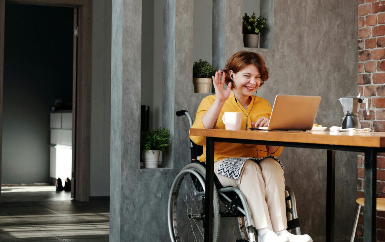 Frau im Rollstuhl arbeitet am Laptop und winkt fröhlich in die Kamera. 